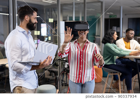 Biracial businesswoman wearing vr headset by caucasian businessman with laptop during meeting Biracial businesswoman wearing vr headset by caucasian businessman with laptop during meeting 90337934