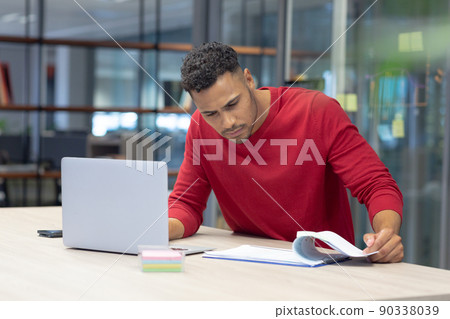 Hispanic male advisor analyzing document while sitting with laptop at modern workplace 90338039