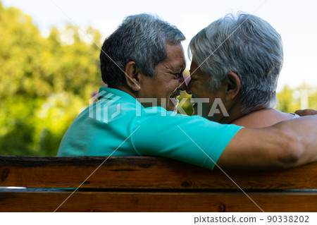 Close-up of smiling biracial senior couple looking at each other while sitting on bench in park Close-up of smiling biracial senior couple looking at each other while sitting on bench in park 90338202