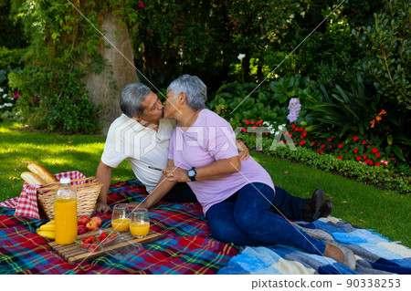 Biracial senior couple kissing while sitting with juice and food on blanket against plants in park 90338253