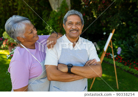 Smiling biracial senior woman with hand on husband's shoulder standing against plants in yard 90338254