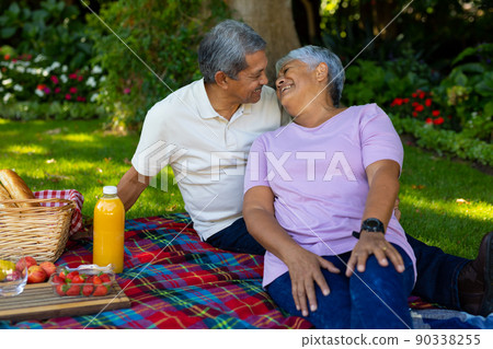 Biracial senior couple looking at each other while sitting with food and drink on blanket in park Biracial senior couple looking at each other while sitting with food and drink on blanket in park 90338255
