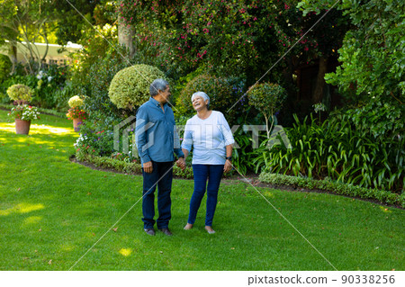 Biracial happy senior woman holding husband's hand while standing against plants and trees in park Biracial happy senior woman holding husband's hand while standing against plants and trees in park 90338256