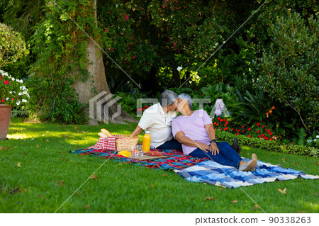 Biracial senior couple kissing while sitting with food and drink on blanket against plants in park Biracial senior couple kissing while sitting with food and drink on blanket against plants in park 90338263