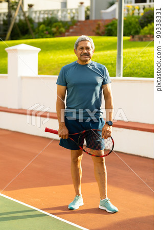 Portrait of confident biracial senior man holding tennis racket while standing in tennis court 90338331