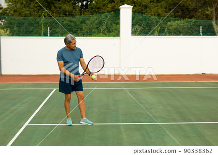 Full length of biracial senior man holding racket and ball playing tennis at tennis court 90338362