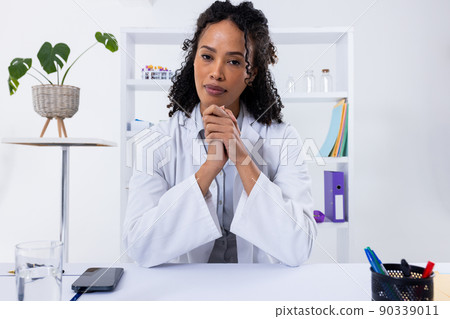Portrait of confident african american mid adult female doctor with hands clasped sitting at desk 90339011