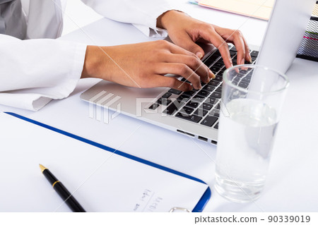 Cropped hands of african american mid adult female doctor tying over laptop by water glass on desk Cropped hands of african american mid adult female doctor tying over laptop by water glass on desk 90339019