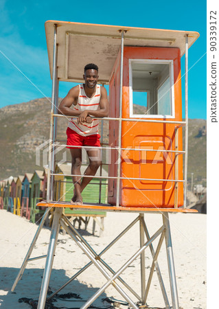 Portrait of smiling african american young man standing by railing on lifeguard hut against blue sky Portrait of smiling african american young man standing by railing on lifeguard hut against blue sky 90339172