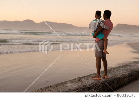 African american young man carrying son while standing on rock at beach against clear sky at sunset African american young man carrying son while standing on rock at beach against clear sky at sunset 90339173