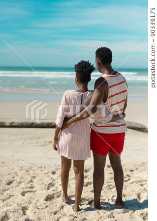 Rear view of african american young couple with arms around looking at sea while standing on beach Rear view of african american young couple with arms around looking at sea while standing on beach 90339175