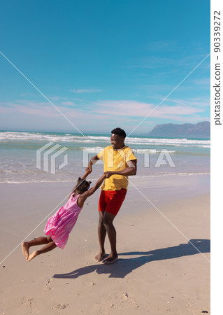 Happy african american young man holding daughter's hands and spinning her at beach against blue sky 90339272