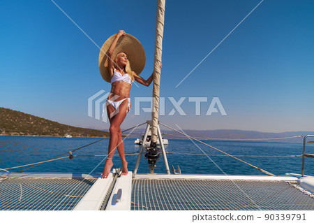 Beautiful young blond woman in bikini standing on catamaran bow at sunny summer day Beautiful young blond woman in bikini standing on catamaran bow at sunny summer day 90339791
