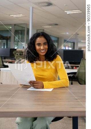 Portrait of smiling multiracial businesswoman with document sitting at desk in modern workplace Portrait of smiling multiracial businesswoman with document sitting at desk in modern workplace 90339905