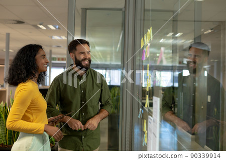Happy multiracial colleagues looking at strategy on glass wall while discussing at modern workplace Happy multiracial colleagues looking at strategy on glass wall while discussing at modern workplace 90339914