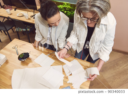 Two diverse stylish women tailors discuss sewing patterns while developing new collection of clothes in fashion design workshop and tailoring atelier Two diverse stylish women tailors discuss sewing patterns while developing new collection of clothes in fashion design workshop and tailoring atelier 90340033