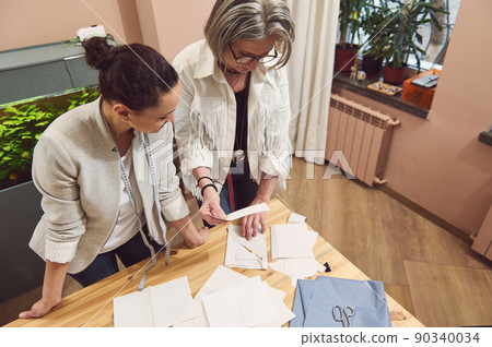 Creative multi-ethnic team of two seamstresses designers work on creation of new seasonal clothing collection, discussing sewing patterns and fabrics in fashion design studio. 90340034