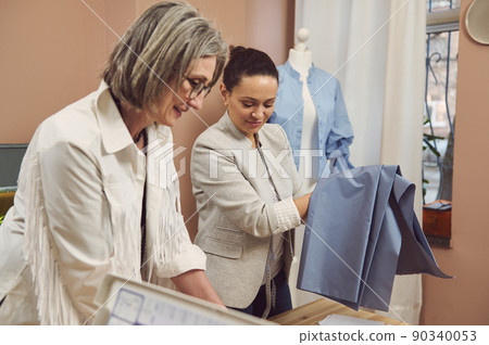 Portrait of a middle-aged Hispanic fashion designer in a stylish jacket holding blue fabric to create a new collection of women's dress shirts in a fashion design workshop Portrait of a middle-aged Hispanic fashion designer in a stylish jacket holding blue fabric to create a new collection of women's dress shirts in a fashion design workshop 90340053