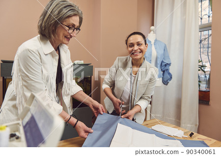 Waist-length portrait of two mature female tailors making clothes patterns on fabric while sewing a classic women's shirt in a fashion design workshop Waist-length portrait of two mature female tailors making clothes patterns on fabric while sewing a classic women's shirt in a fashion design workshop 90340061
