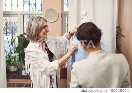 Charming mature female fashion designer and her assistant try on new shirt on mannequin, prepare a new collection of classic clothes for exhibition in fashion design studio 90340064