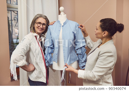 Senior fashion designer smiles posing with a dummy wearing blue shirt from new collection while her colleague tailor takes measurements and works on improvement details of new garment 90340074