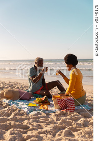 African american couple having juice while sitting with food on blanket at beach against clear sky African american couple having juice while sitting with food on blanket at beach against clear sky 90340179