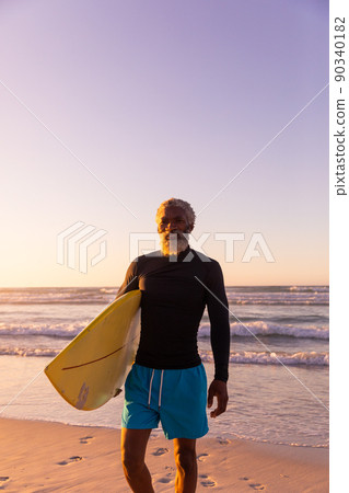 Bearded african american senior man with surfboard standing against sea and clear sky at sunset 90340182