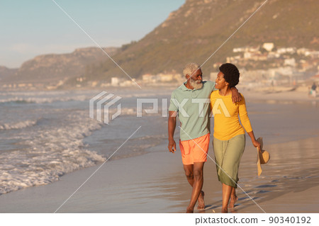 African american senior man with hand on mature woman's shoulder walking at beach at sunset 90340192