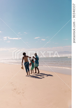 African american grandparents holding grandson's hands and walking at sandy beach against blue sky 90340337