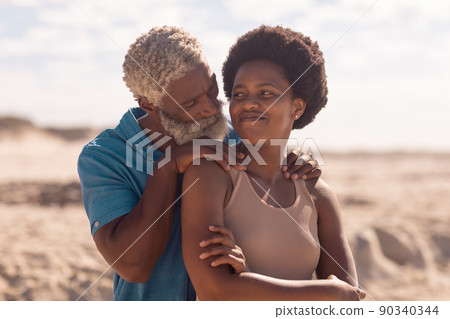 African american bearded senior man with hands on mature woman's shoulders at beach against sky 90340344