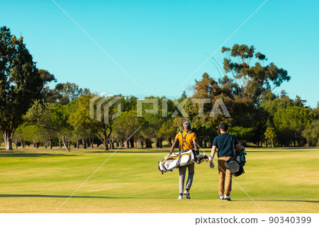Rear view of multiracial young male friends with golf bags walking at golf course against clear sky Rear view of multiracial young male friends with golf bags walking at golf course against clear sky 90340399