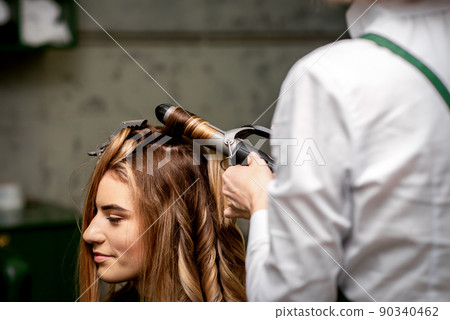 The female hairdresser is curling hair for a brown-haired young caucasian woman in a beauty salon 90340462