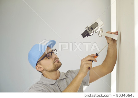 Concentrated young male technician repairs or installs CCTV system on wall inside building. Concentrated young male technician repairs or installs CCTV system on wall inside building. 90340643