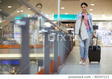Cheerful attractive young lady with coat on arm carrying wheeled luggage and using smartphone while hurrying for boarding onto airplane 90347221