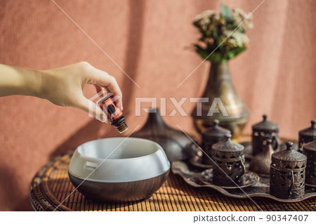 Woman adding essential oil to aroma diffuser on table 90347407