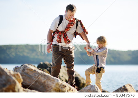 Cute schoolchild and his mature father hiking together on mountain near sea and exploring nature. 90347576