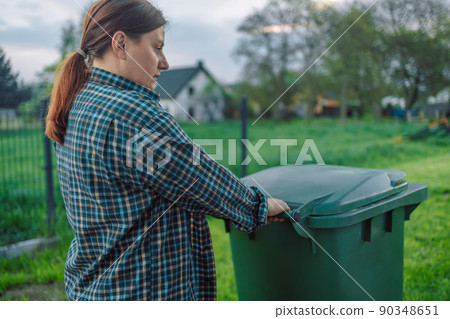 European 20s girl take out the trash can sorting garbage near a home. Female transports a green container with garbage to the backyard European 20s girl take out the trash can sorting garbage near a home. Female transports a green container with garbage to the backyard 90348651