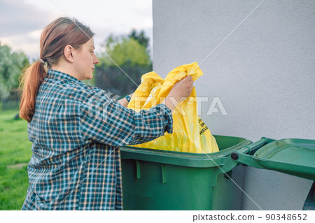 European 30s woman throwing garbage into the recycling bin in the backyard near the house. Separating recyclable garbage plastic  90348652
