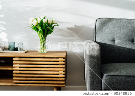 Scandinavian home interior with spring bouquet of white tulip flowers in glass vase, tray with candles standing on a wooden cabinet. Minimalist design with gray sofa and white wall. Springtime. 90348936