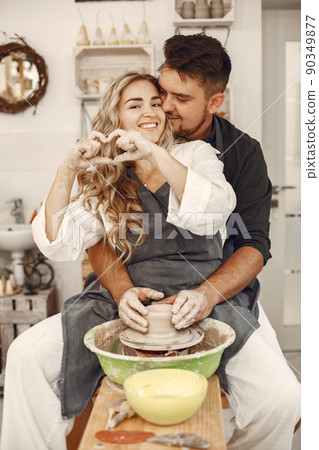 Mutual creative work. Young beautiful couple in casual clothes and aprons. People creating a bowl on a pottery wheel in a clay studio. Mutual creative work. Young beautiful couple in casual clothes and aprons. People creating a bowl on a pottery wheel in a clay studio. 90349877