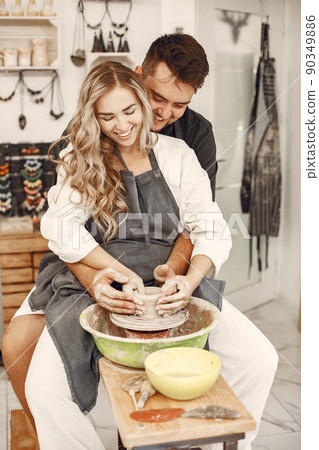 Mutual creative work. Young beautiful couple in casual clothes and aprons. People creating a bowl on a pottery wheel in a clay studio. Mutual creative work. Young beautiful couple in casual clothes and aprons. People creating a bowl on a pottery wheel in a clay studio. 90349886