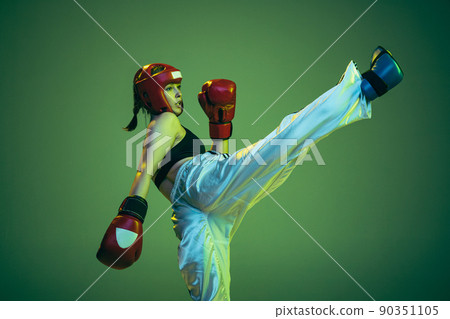 Leg kick. Flexible female boxer in boxing gloves and helmet practicing isolated on green studio background in neon light. Sport, competition, challenges, results, success concept Leg kick. Flexible female boxer in boxing gloves and helmet practicing isolated on green studio background in neon light. Sport, competition, challenges, results, success concept 90351105