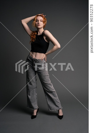 Confident young woman standing in gray studio with hand on waist 90352278