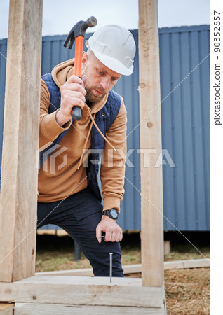 Man worker building wooden frame house on pile foundation. Carpenter hammering nail into wooden board, using hammer. Carpentry concept. 90352857