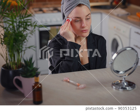 young woman looks in mirror using facial gua sha jade board at home. 90353125