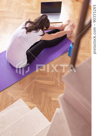 Young happy caucasian woman stretching in the living room while looking at her laptop. Health care and wellness at home concept. Young happy caucasian woman stretching in the living room while looking at her laptop. Health care and wellness at home concept. 90355265