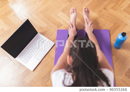 Young happy caucasian woman stretching in the living room while looking at her laptop. Health care and wellness at home concept. Young happy caucasian woman stretching in the living room while looking at her laptop. Health care and wellness at home concept. 90355268