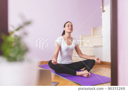Young happy caucasian woman meditating in the living room while looking at her laptop. Health care and wellness at home concept. Young happy caucasian woman meditating in the living room while looking at her laptop. Health care and wellness at home concept. 90355269