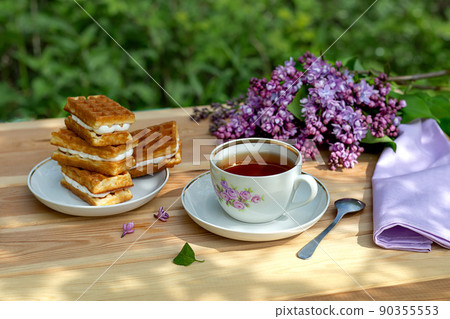 Tea party with Viennese waffles and a branch of lilac on a wooden table with shadows against the background of the garden on a sunny summer day. Tea party with Viennese waffles and a branch of lilac on a wooden table with shadows against the background of the garden on a sunny summer day. 90355553