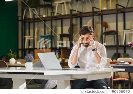 Front view of tired architect, designer sitting at table in furniture shopping. Handsome brunette with beard looking down, holding head by hands, thinking. Concept of shopping. Front view of tired architect, designer sitting at table in furniture shopping. Handsome brunette with beard looking down, holding head by hands, thinking. Concept of shopping. 90356970